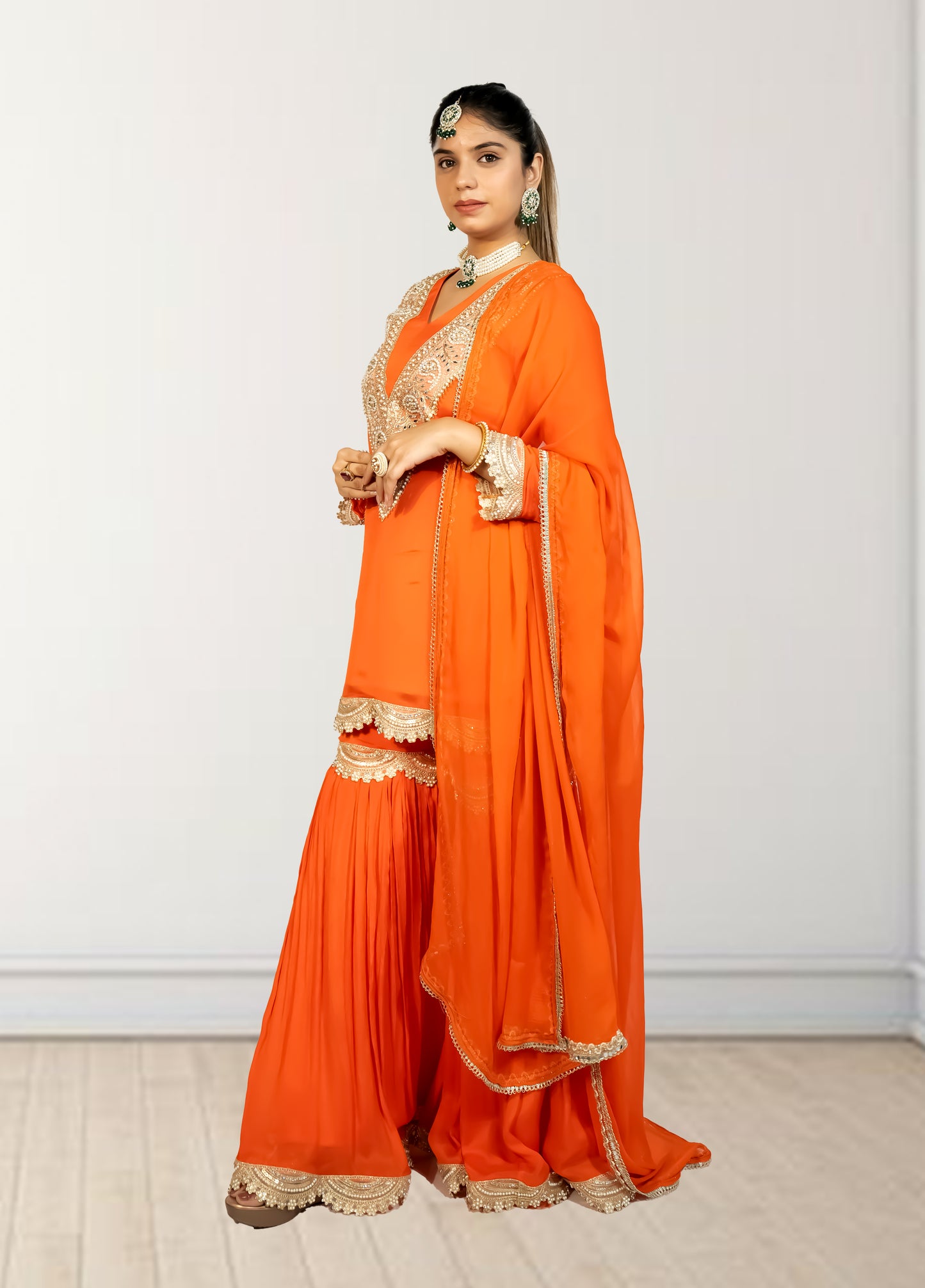 Woman wearing an orange saree with gold embroidery against a white wall.