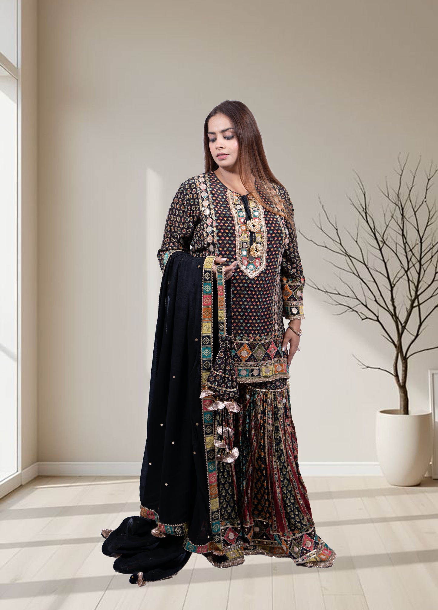 Woman wearing a patterned dress with a black dupatta in a room with a large window and a plant.