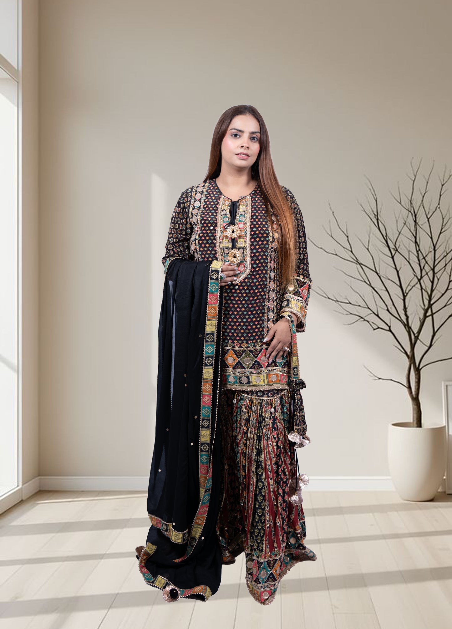 Woman wearing a traditional outfit with a patterned top and black dupatta in a minimalistic room.