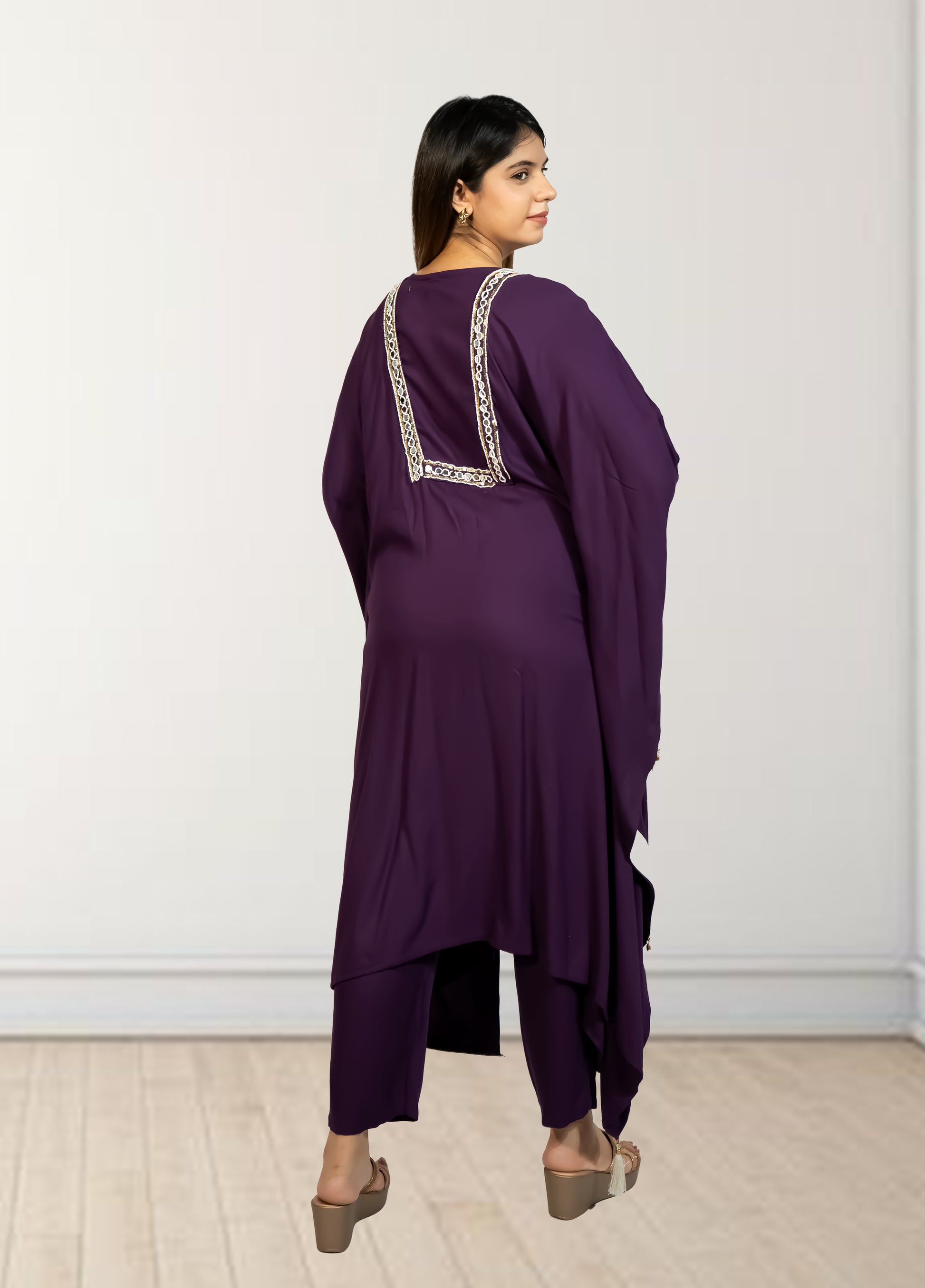 Woman wearing a purple outfit with intricate detailing on the back against a white wall.