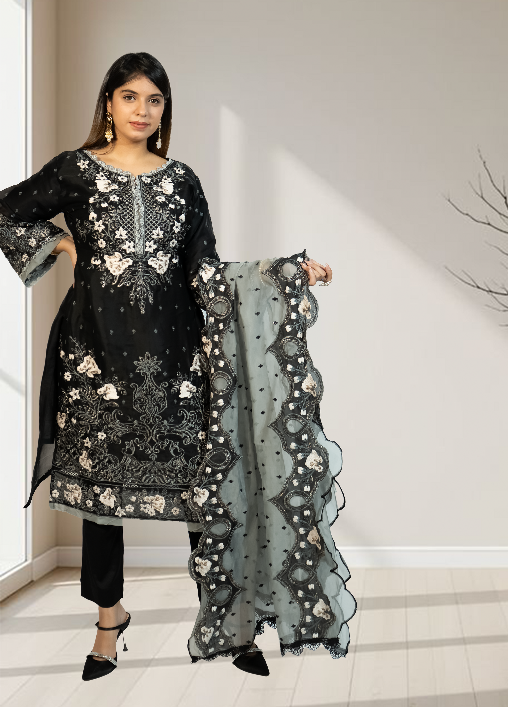 Woman holding a black embroidered outfit with a matching dupatta in a minimalistic setting