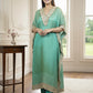 Woman in a teal traditional outfit standing in a well-lit room with large windows.