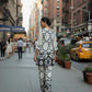 Woman in a black and white patterned outfit walking on a city street with buildings and a taxi in the background.