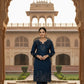 Woman in a blue traditional outfit standing in front of an architectural archway with a building in the background.
