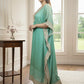 Woman in a light green saree standing in a room with large windows.