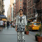 Woman in a black and white patterned outfit standing on a city street with a yellow taxi in the background.