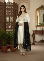 Woman in a white and black traditional outfit standing in a decorated room with plants and a mirror.