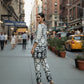 Woman in a black and white patterned outfit walking on a city street with buildings and a taxi in the background.