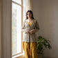 Woman in a patterned cardigan and yellow pants standing by a window with a plant in the background.