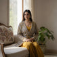 Woman in a patterned cardigan and yellow pants sitting on a couch by a window.