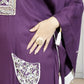 Purple garment with white embroidery and a hand wearing a ring, on a neutral background
