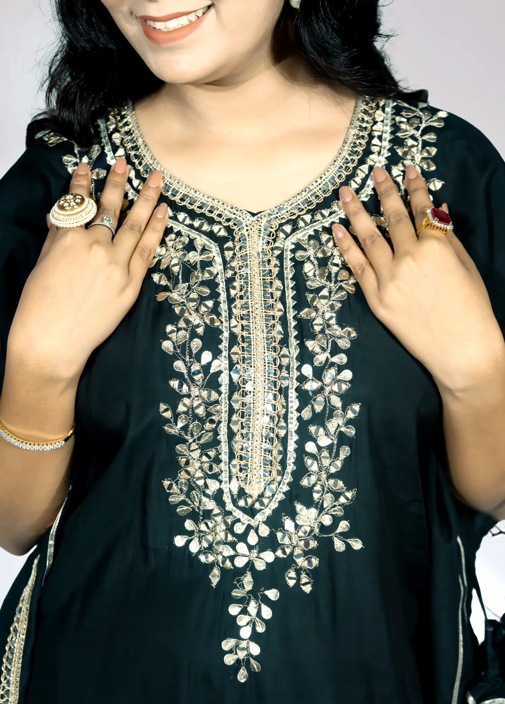 Woman wearing a black dress with intricate white embroidery, holding up her hands to show off jewelry.