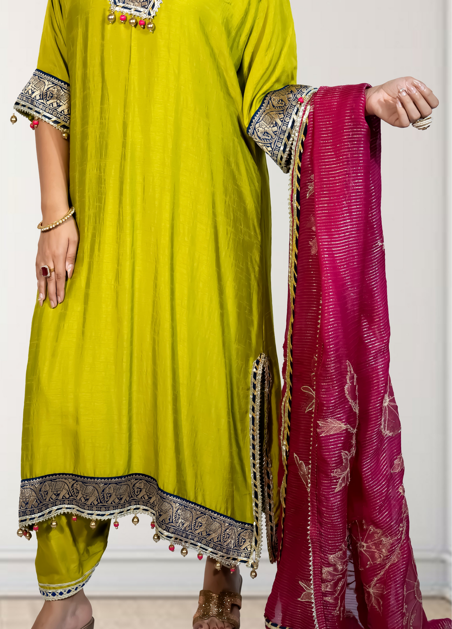 Person wearing a green traditional outfit with a pink dupatta, standing against a white wall.