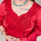 Person wearing a red embroidered outfit with a green necklace against a neutral background