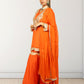 Woman wearing an orange saree with gold embroidery against a white wall.