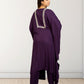 Woman wearing a purple outfit with intricate detailing on the back against a white wall.