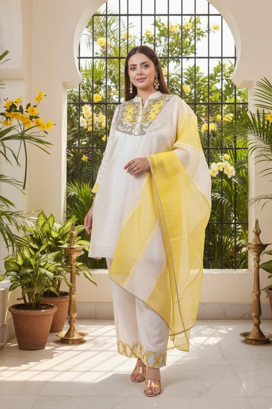Woman in a white and yellow traditional outfit standing in a well-lit room with plants and a window.