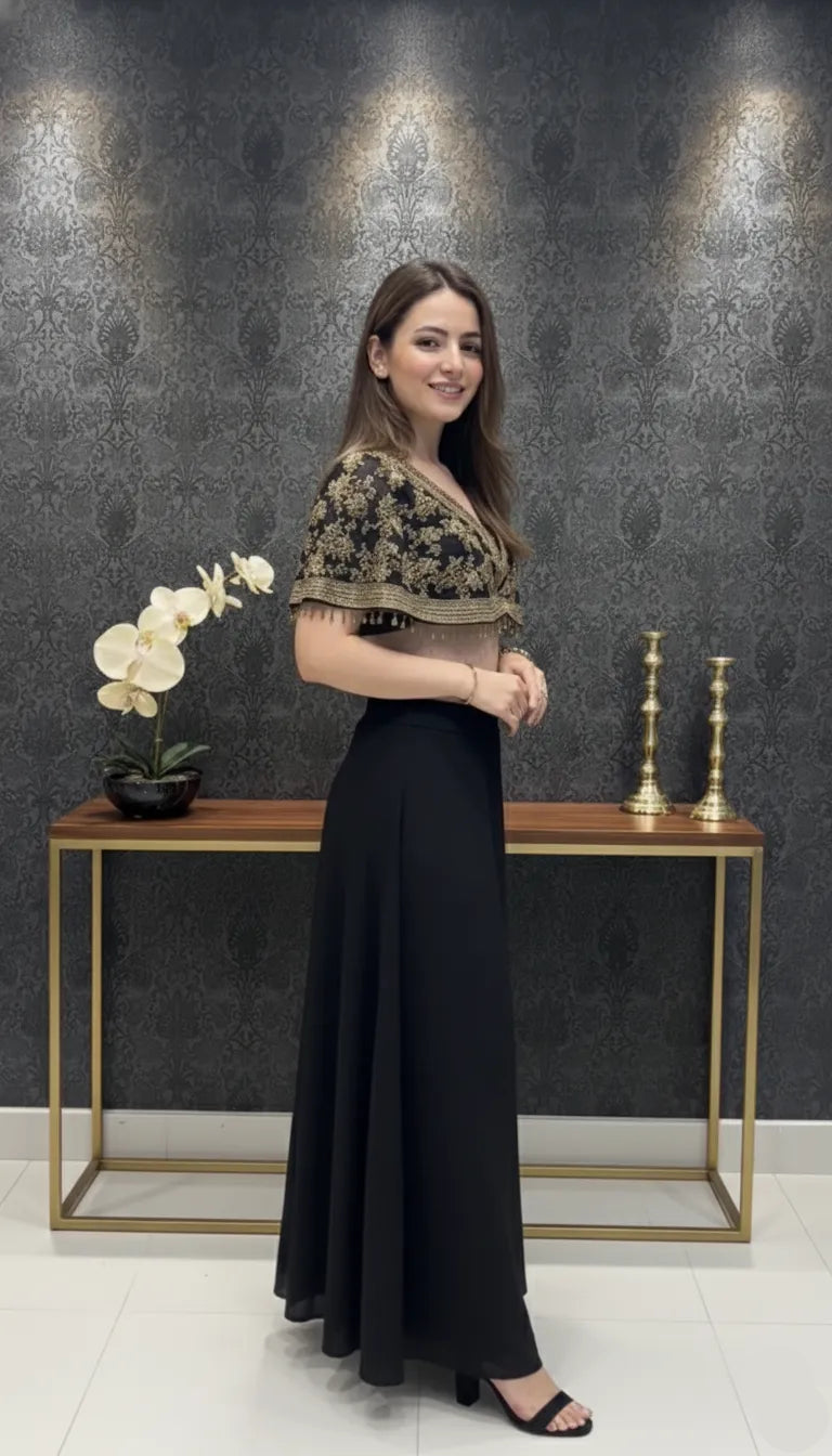 Woman in a black dress with floral top standing next to a decorative table against a dark wall.