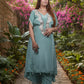 Woman in a teal traditional outfit standing in a garden with pink flowers and stone pathway.