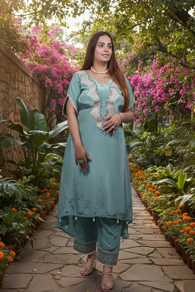 Woman in a teal traditional outfit standing in a garden with pink flowers and stone pathway.