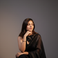 Woman in a black saree sitting against a gray background