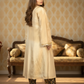 Woman in a beige traditional outfit standing in a room with patterned wallpaper and furniture.