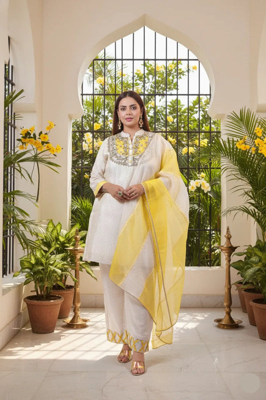 Woman in a white and yellow traditional outfit standing in a sunlit room with plants.
