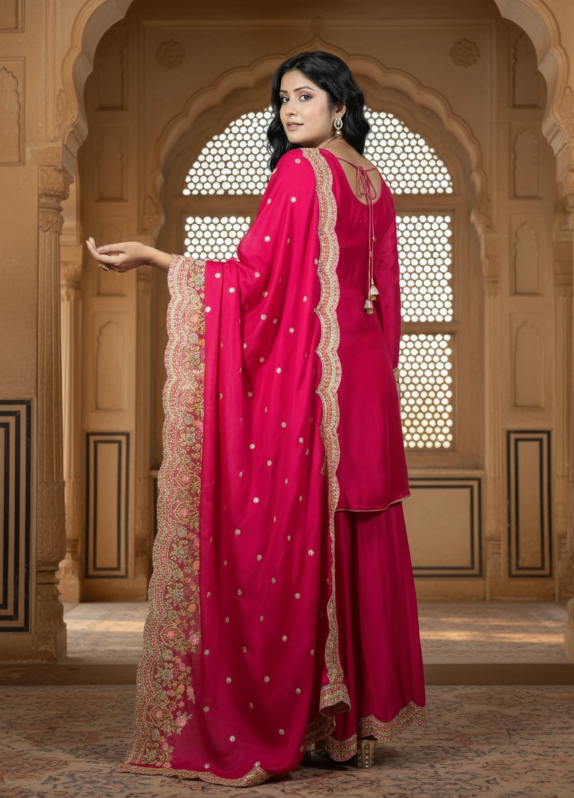 Woman in a bright pink traditional outfit with gold details standing in front of an ornate archway.
