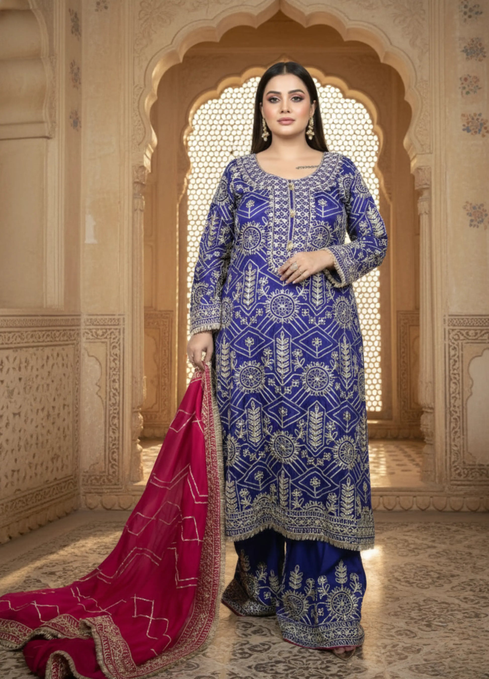 Woman in a blue embroidered traditional outfit with a red dupatta in an ornate architectural setting.