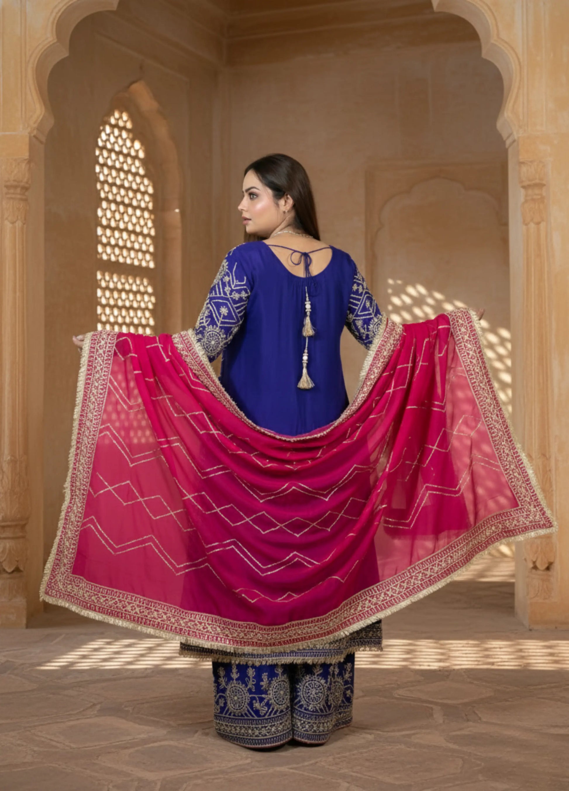 Woman in a blue and pink traditional outfit standing in a decorative interior setting.