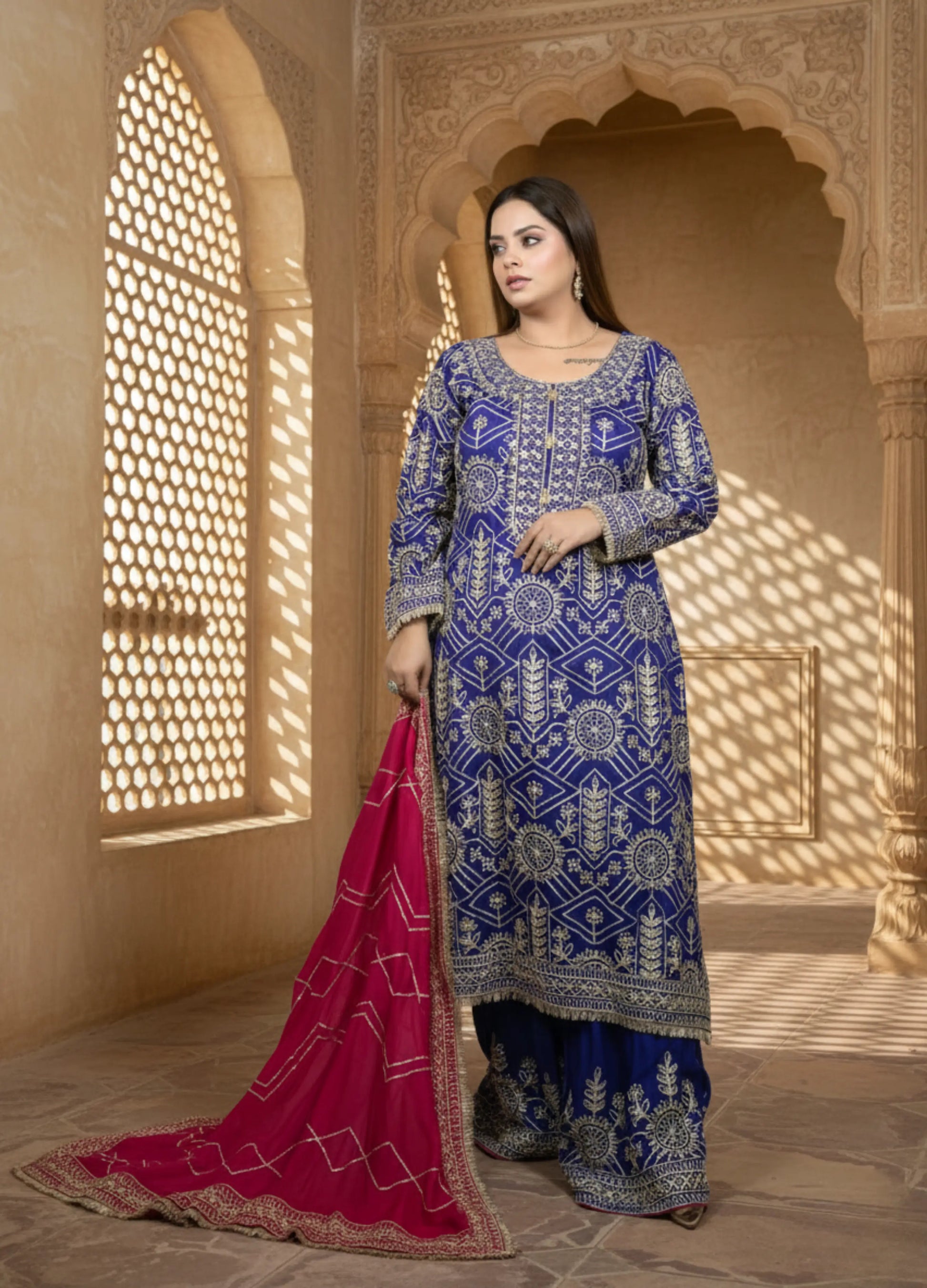 Woman in a blue embroidered traditional outfit with a pink dupatta in a sunlit room.