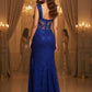 Woman in a blue lace evening gown standing in a grand room with chandeliers.