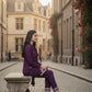 Woman in a purple outfit sitting on a stone bench in an urban setting with buildings and flowers in the background.