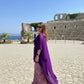 Woman in a purple dress standing in front of ancient ruins with blue sky and ocean in the background