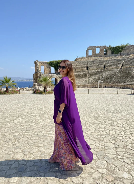 Woman in a purple dress standing in front of ancient ruins with blue sky and ocean in the background
