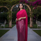 Woman in a pink saree with a sequined skirt standing in an outdoor setting with greenery and arches.