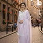 Woman in a light purple traditional outfit standing on a cobbled street with buildings and people in the background.