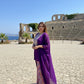 Woman in a purple dress standing in front of ancient ruins with blue sky and ocean in the background